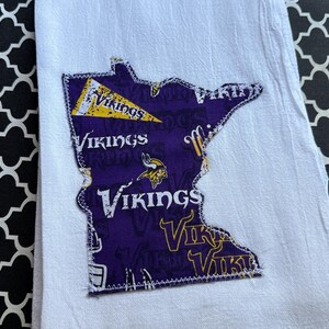 May include: White hand towel with a purple patch in the shape of Minnesota. The patch features the word "Vikings" in white and gold, along with a Viking ship logo. The towel is on a black and white patterned surface.