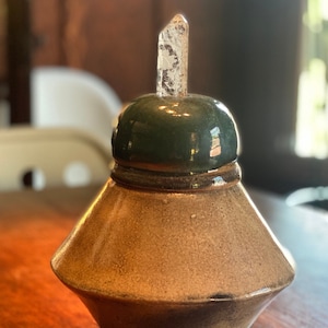 Pode incluir: Um jarro de cerâmica artesanal com tampa verde escuro e um cristal transparente. O jarro tem uma forma única com um acabamento texturizado castanho claro. Provavelmente é usado para armazenar especiarias ou outros itens pequenos.