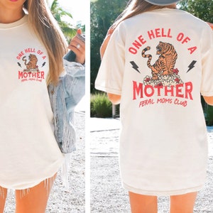 May include: A white t-shirt with a graphic of a tiger and the text "One Hell of a Mother Feral Moms Club".
