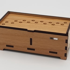 Puede incluir: Una caja de madera con un borde marrón oscuro y una tapa con un patrón de huellas de patas. La caja está hecha de madera clara y tiene un diseño simple.