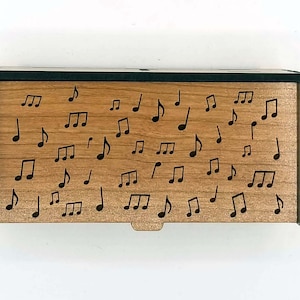 May include: A wooden box with a light brown finish and black trim. The box is decorated with numerous black musical notes. The design is simple and elegant, suitable for storing small items or as a decorative piece.