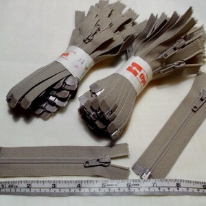 May include: A close-up of a pile of beige zippers with silver sliders. The zippers are bundled together and labeled "13" and "50". A ruler is in the bottom of the image.