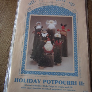 Könnte beinhalten: Eine Packung mit Schnittmustern für Holiday Potpourri II, die sechs festliche Roly Poly-Figuren zeigt, darunter Weihnachtsmann und Engel, auf Holzblöcken. Die Verpackung hat blaue und weiße dekorative Elemente und den Text "all. cooped up."