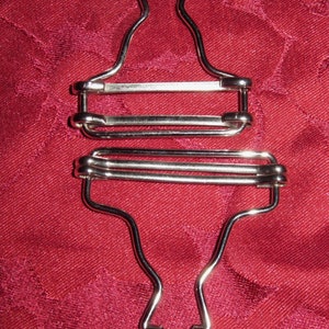 Könnte beinhalten: Ein silberner Hosenträgerclip mit Haken- und Schlaufen-Design. Der Clip hat drei verstellbare Schlaufen zum Befestigen an der Kleidung.