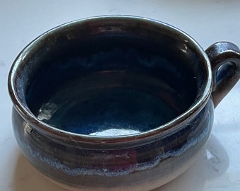May include: A dark blue ceramic bowl with a single handle. The bowl has a glazed finish and a light brown rim.