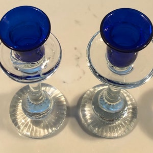 May include: Two clear glass candle holders with blue glass candle cups. The candle holders have a fluted base and a clear glass stem.