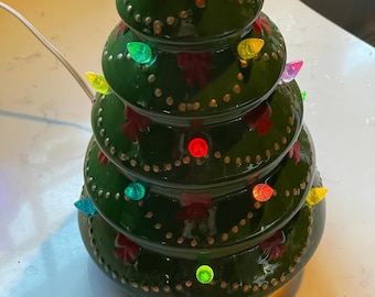 May include: A ceramic Christmas tree with a green glaze, adorned with colorful lights and red bow accents. The tree has a gold-colored top and is lit with multi-colored bulbs. A white power cord extends from the base.