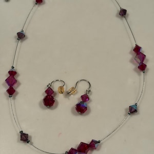 May include: A jewelry set consisting of a necklace and a pair of earrings. The necklace features a thin silver-toned wire strand accented with faceted, translucent red and purple beads. The matching earrings feature similar red beads on silver-toned hooks.