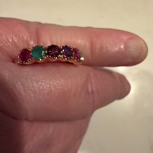 May include: A gold ring featuring five round, multi-coloured gemstones set in a row. The stones include a pink, a green, a deep red, a purple, and a lighter pink or clear stone, all held in place by small gold prongs along the band of the ring.