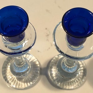 May include: Two clear glass candle holders with blue glass candle cups. The candle holders have a fluted base and a clear glass stem.