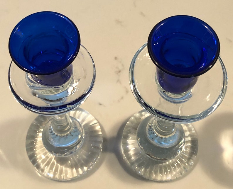 May include: Two clear glass candle holders with blue glass candle cups. The candle holders have a fluted base and a clear glass stem.