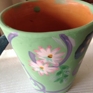 May include: A hand-painted, light green flower pot with a flared shape. The pot features two white daisies with pink tips and yellow centers, along with purple and green painted accents. The interior is a natural terracotta color.