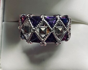 May include: A wide silver-toned band ring featuring a repeating diamond-shaped pattern. The design alternates between deep purple triangular stones and small, square, dark metallic studs set within a textured, intricate silver metal framework.