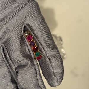 May include: A gold band ring featuring a row of six round, multi-coloured gemstones including clear, pink, purple, red, green, and magenta stones set in a scalloped metal frame. The ring is held against a smooth, grey fabric surface.