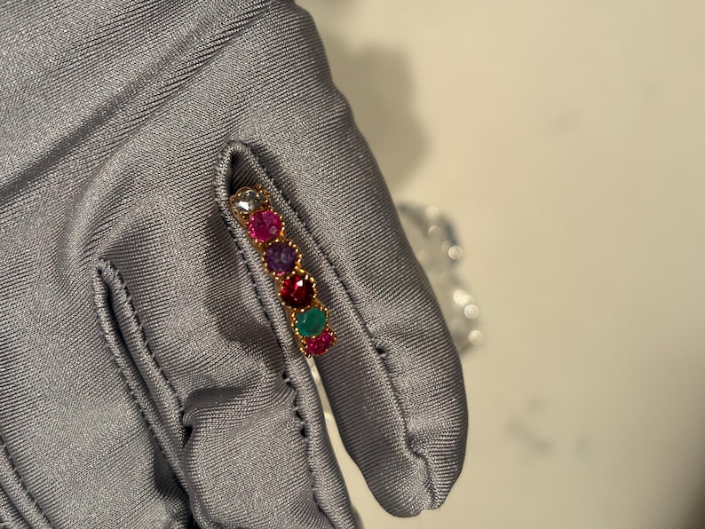 May include: A gold band ring featuring a row of six round, multi-coloured gemstones including clear, pink, purple, red, green, and magenta stones set in a scalloped metal frame. The ring is held against a smooth, grey fabric surface.