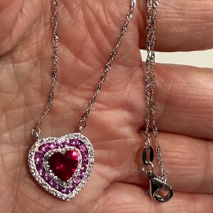 May include: A silver-toned necklace with a heart-shaped pendant. The pendant features a central red gemstone, surrounded by a layer of pink stones, and an outer layer of clear stones. The chain is delicate and has a lobster clasp.