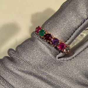 May include: A gold band ring featuring a row of six round-cut gemstones in various colours, including red, green, purple, and clear stones, set in a prong setting. The ring is displayed on a smooth, light grey fabric surface with visible texture.