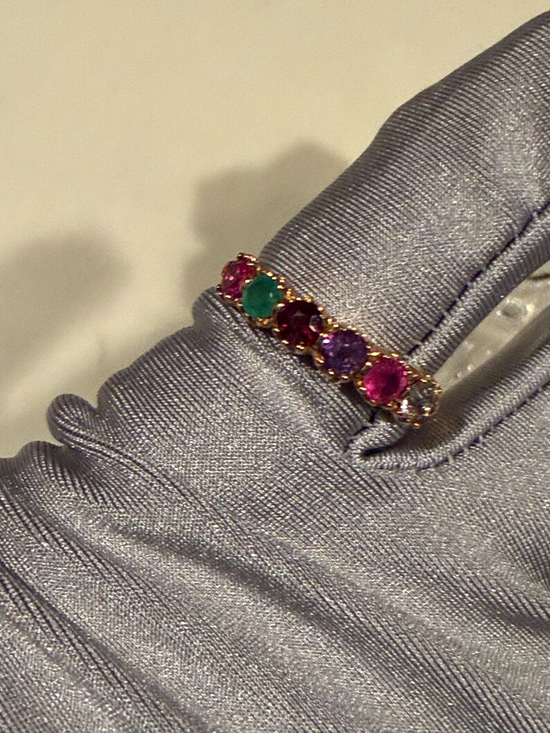 May include: A gold band ring featuring a row of six round-cut gemstones in various colours, including red, green, purple, and clear stones, set in a prong setting. The ring is displayed on a smooth, light grey fabric surface with visible texture.