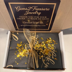 May include: A cardboard shipping box containing a package wrapped in black paper with gold script, tied with a gold sheer ribbon and topped with gold foil flakes. The box lid features a black label with gold text reading Gems2Treasure Jewelry.