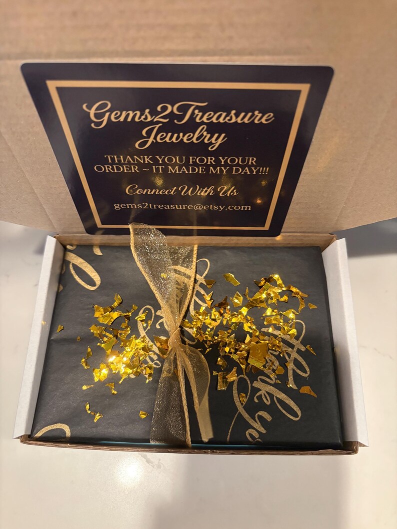 May include: A cardboard shipping box containing a package wrapped in black paper with gold script, tied with a gold sheer ribbon and topped with gold foil flakes. The box lid features a black label with gold text reading Gems2Treasure Jewelry.