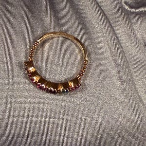 May include: A circular ring featuring a metallic band with a beaded texture along the upper edge. The lower portion of the ring is set with a row of small, vibrant, multi-coloured gemstones including shades of pink, purple, and blue in a prong setting.