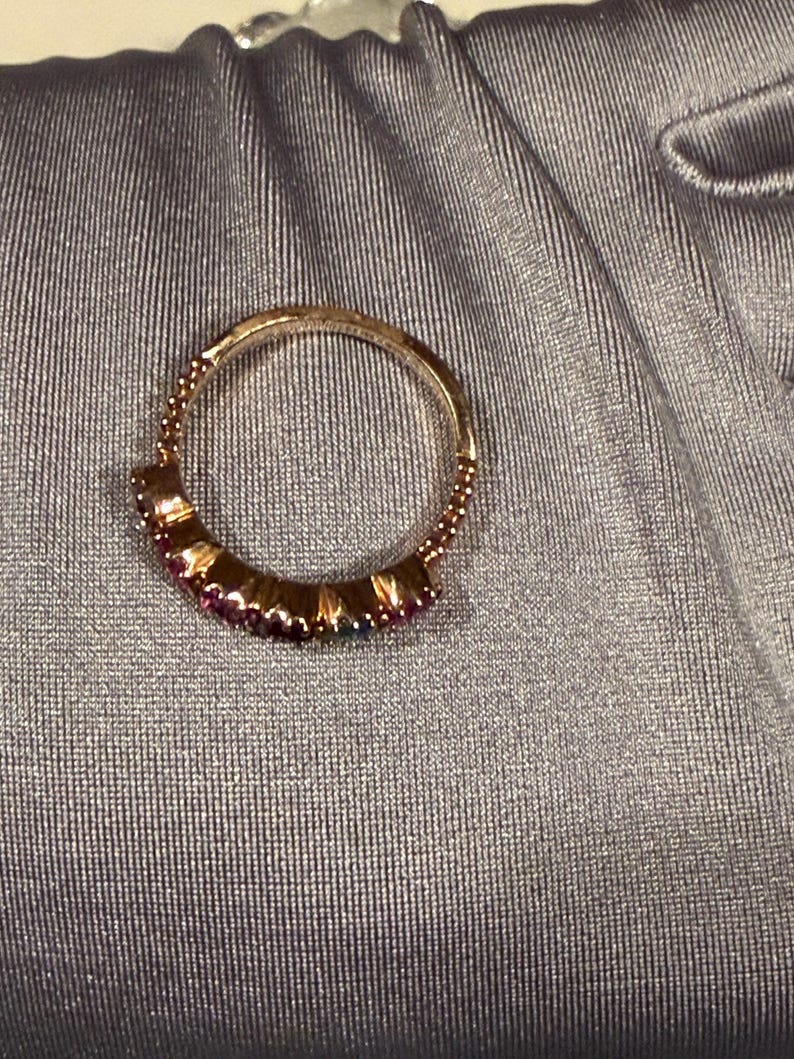 May include: A circular ring featuring a metallic band with a beaded texture along the upper edge. The lower portion of the ring is set with a row of small, vibrant, multi-coloured gemstones including shades of pink, purple, and blue in a prong setting.
