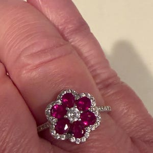 May include: A flower-shaped ring featuring a central round diamond surrounded by six ruby gemstones. The ruby petals are encircled by a halo of smaller diamonds, all set in a silver band. The ring is a piece of jewelry.