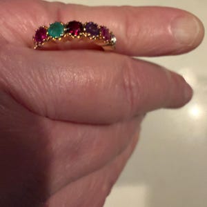 May include: A gold band ring featuring a row of six round-cut gemstones in various colours, including red, green, deep pink, purple, and a clear stone, all set in a prong setting along the top of the band.