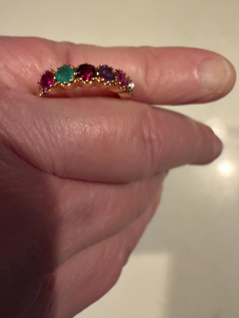 May include: A gold band ring featuring a row of six round-cut gemstones in various colours, including red, green, deep pink, purple, and a clear stone, all set in a prong setting along the top of the band.