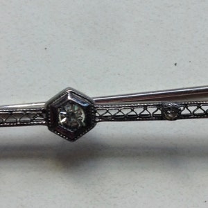 May include: A silver bar pin with a central hexagonal stone and smaller stones on either side. The pin is decorated with a delicate filigree design.