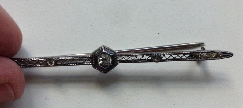 May include: A silver bar pin with a central hexagonal stone and smaller stones on either side. The pin is decorated with a delicate filigree design.