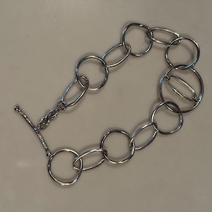 May include: Silver-toned chain bracelet featuring a toggle clasp. The bracelet is composed of various sized oval and circular links. The links are connected to form a flexible chain. The bracelet is displayed on a neutral surface.