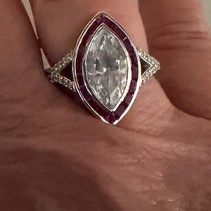 May include: A silver ring featuring a large, marquise-cut, clear gemstone at the center, framed by a row of small, rectangular, red gemstones. The ring's band is adorned with small, clear gemstones on each side of the marquise-cut center stone.