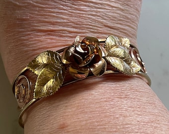 May include: A gold-toned metal cuff bracelet featuring a central three-dimensional rose flower ornament flanked by two textured gold leaves. The band of the bracelet is constructed from thin, polished gold wire that forms decorative spiral patterns.