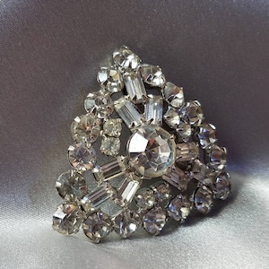May include: A silver tone brooch with a triangular shape, featuring a cluster of clear rhinestones and a large, central, faceted rhinestone.