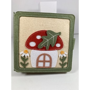 May include: A square, felt book with a green border and a beige background. The cover features a red mushroom with white spots, a green leaf, and a white door. Two white daisies with yellow centers flank the mushroom.