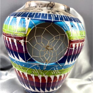 May include: A ceramic vase featuring a circular cutout covered with a woven web pattern. The exterior is decorated with horizontal bands of blue, green, purple, and red, accented by white vertical lines and a crackled texture on the upper rim.