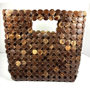 May include: A rectangular handbag constructed from numerous small, circular, dark brown coconut shell discs. The discs are arranged in a grid pattern and linked together, forming the body of the bag. It features a rectangular cutout handle at the top.