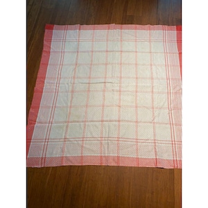 May include: A white and red checkered tablecloth. The tablecloth has a white center with a grid pattern and a red border. The red border is approximately 2 inches wide. The tablecloth is folded and lying on a wooden surface.