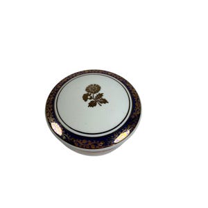 May include: A round, white ceramic trinket box with a gold floral design on the lid. The lid has a dark blue border with gold filigree. The box is a decorative item.