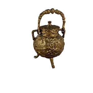 May include: A small, antique brass cauldron with a lid and handle. The cauldron has three small legs and is adorned with intricate embossed designs. The handle is curved and textured, and the lid has a decorative knob. The overall design is ornate and detailed.