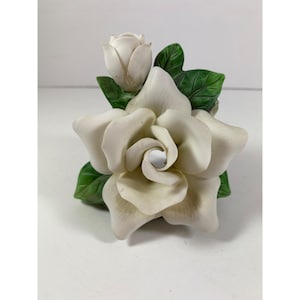 Peut inclure: Une sculpture de fleur de gardénia en céramique avec une grande fleur ouverte et un petit bouton. La fleur est blanc crème avec des pétales détaillés, entourée de feuilles vertes. Un accent floral décoratif.