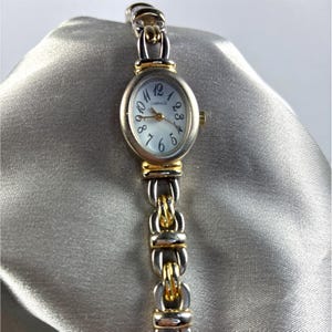 Puede incluir: Un reloj de pulsera plateado y dorado con una esfera ovalada y una correa de eslabones. La esfera del reloj es blanca con números negros y la palabra "CARRIAGE". La correa tiene una combinación de eslabones plateados y dorados.
