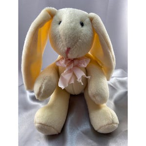 Puede incluir: Un conejito de peluche de color crema con orejas largas y caídas y un lazo rosa. El conejito está sentado sobre una superficie blanca.