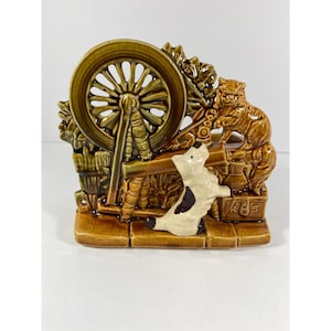 May include: A ceramic figurine depicting a spinning wheel scene with a brown cat and a white and black dog. The figurine is primarily brown and tan with green accents. The base is a brick-like design.