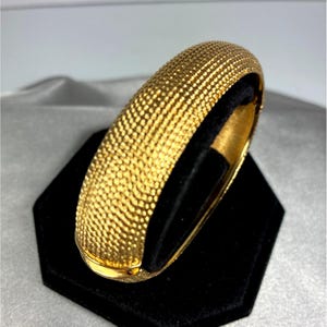 May include: A gold-toned hinged bangle bracelet featuring a textured, granular surface across the exterior. The bracelet has a rounded, domed profile and a polished interior finish, showcasing a secure clasp mechanism visible at the bottom edge.