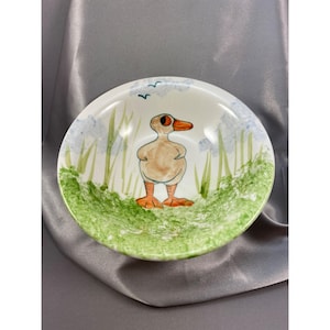Puede incluir: Un cuenco de cerámica blanco con una ilustración de pato pintada a mano. El pato tiene pico y patas naranjas, de pie en una zona de hierba verde. Flores azules y pájaros pintados alrededor del borde.