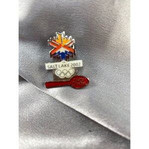 May include: A silver-toned pin featuring the Salt Lake 2002 Olympic logo. The pin includes a snowflake design with orange, yellow, and blue accents, the text "SALT LAKE 2002", the Olympic rings, and a red spoon.
