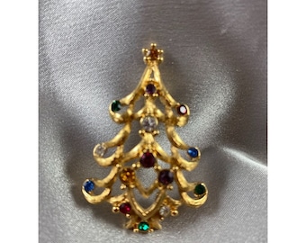 May include: A gold-tone Christmas tree brooch with colorful rhinestones. The tree is decorated with red, green, blue, purple, and clear rhinestones.