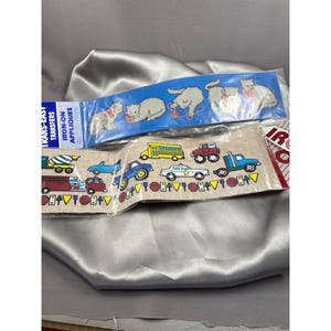 Op de afbeelding: Twee pakketten strijk-applicaties. Eén pakket heeft een blauwe achtergrond met witte katten en de tekst "Iron-On Appliques". Het andere pakket heeft een bruine achtergrond met een rij kleurrijke voertuigen en de tekst "Trans-Easy Transfers".